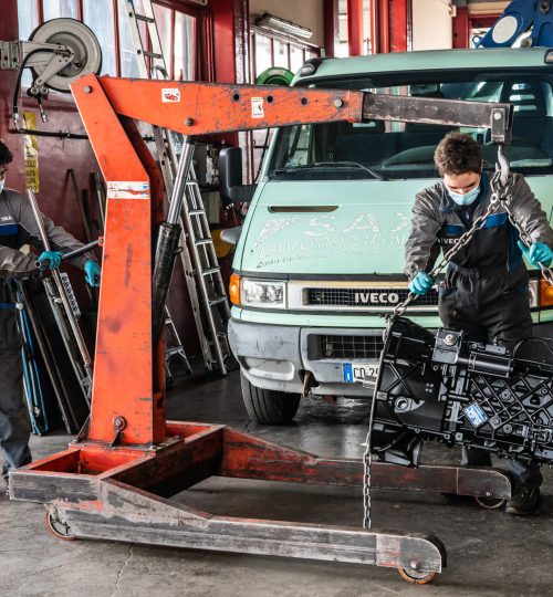 Due meccanici sollevano un motore con una gru.