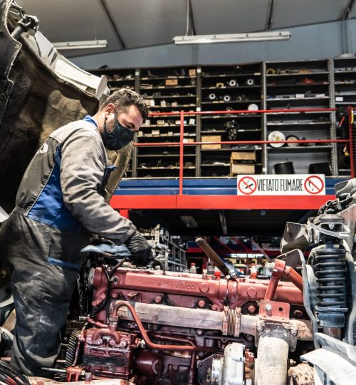 Meccanico lavora su motore in officina, scaffali sullo sfondo.